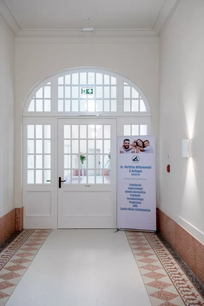 Loepraxis Dr Wilamowskiweb013 Dr Matthias Willamowski Kollegen White interior doorway with arched transom windows and grid panes a tall dental clinic banner stands to the right of the door
