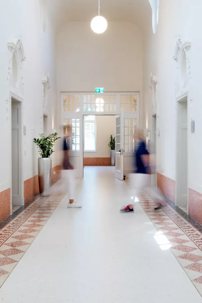 Loepraxis Dr Wilamowskiweb019 Dr Matthias Willamowski Kollegen Bright spacious hallway with white walls decorative moldings and patterned tile trim along the floor blurred people walk toward a doorway at the end