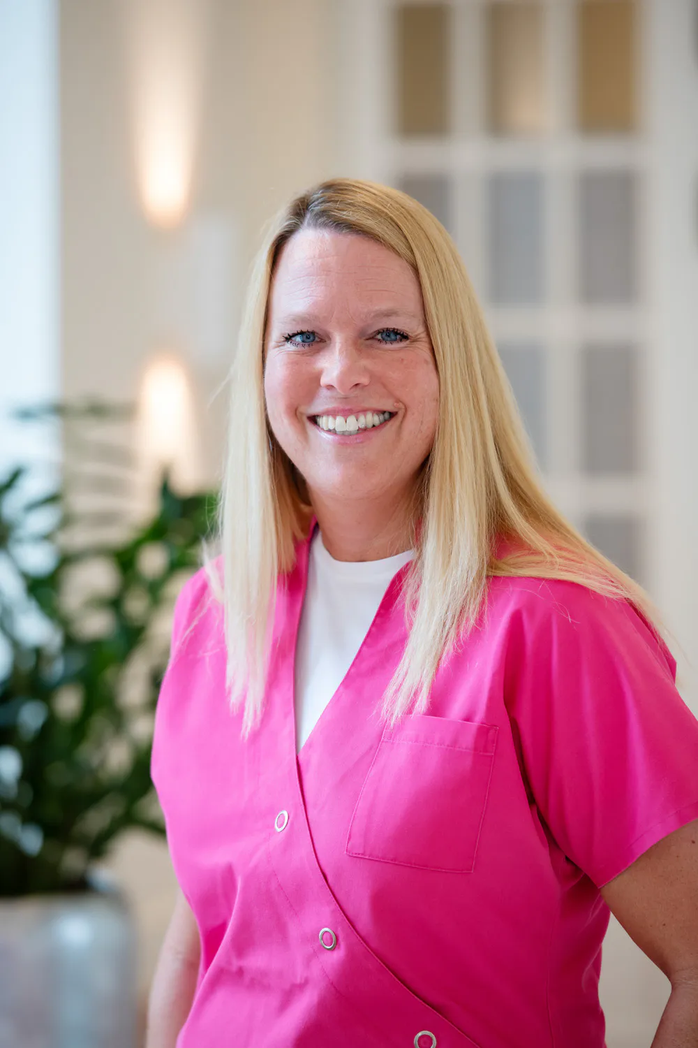 Loepraxis Dr Wilamowskiweb030 Dr Matthias Willamowski Kollegen Smiling woman with long blonde hair wearing bright pink scrubs in a light plant filled indoor space