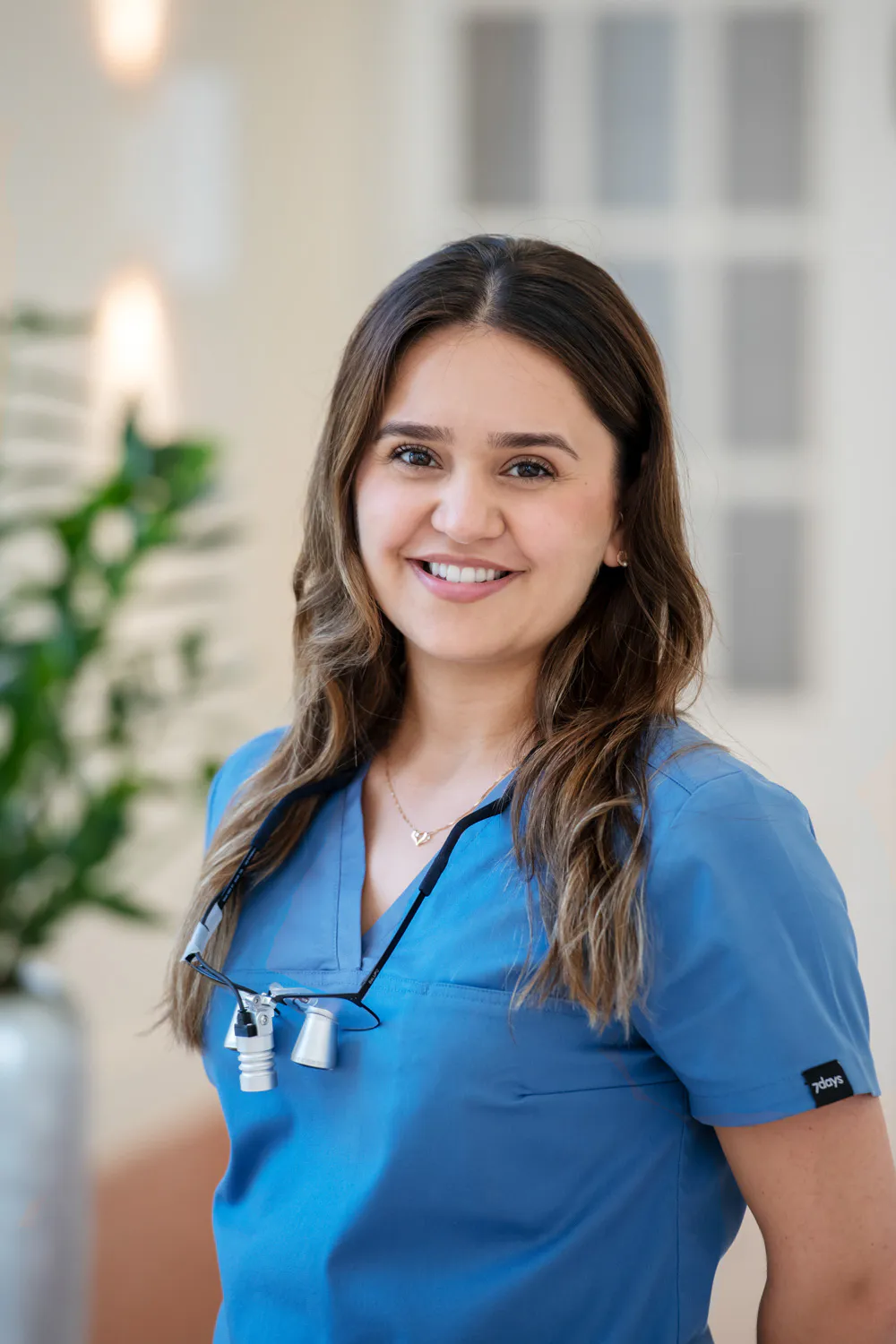Loepraxis Dr Wilamowskiweb033 Dr Matthias Willamowski Kollegen Smiling woman in blue scrubs wearing dental loupes around her neck in a bright clinical setting
