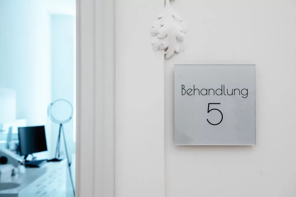 Loepraxis Dr Wilamowskiweb035 Dr Matthias Willamowski Kollegen Door plaque reading Behandlung 5 Treatment 5 on a white wall with a blurred clinic reception area in the background