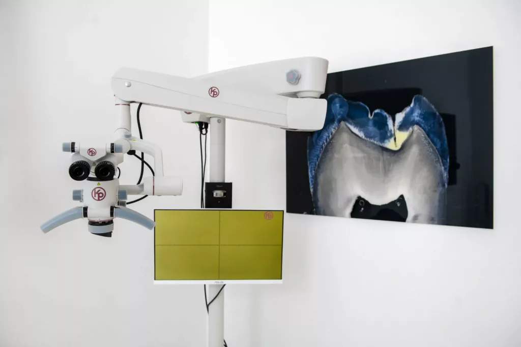 Loepraxis Dr Wilamowskiweb042 Dr Matthias Willamowski Kollegen Dental operating microscope mounted on a stand beside a yellow monitor in a dental clinic setting with a large tooth image on the wall behind it