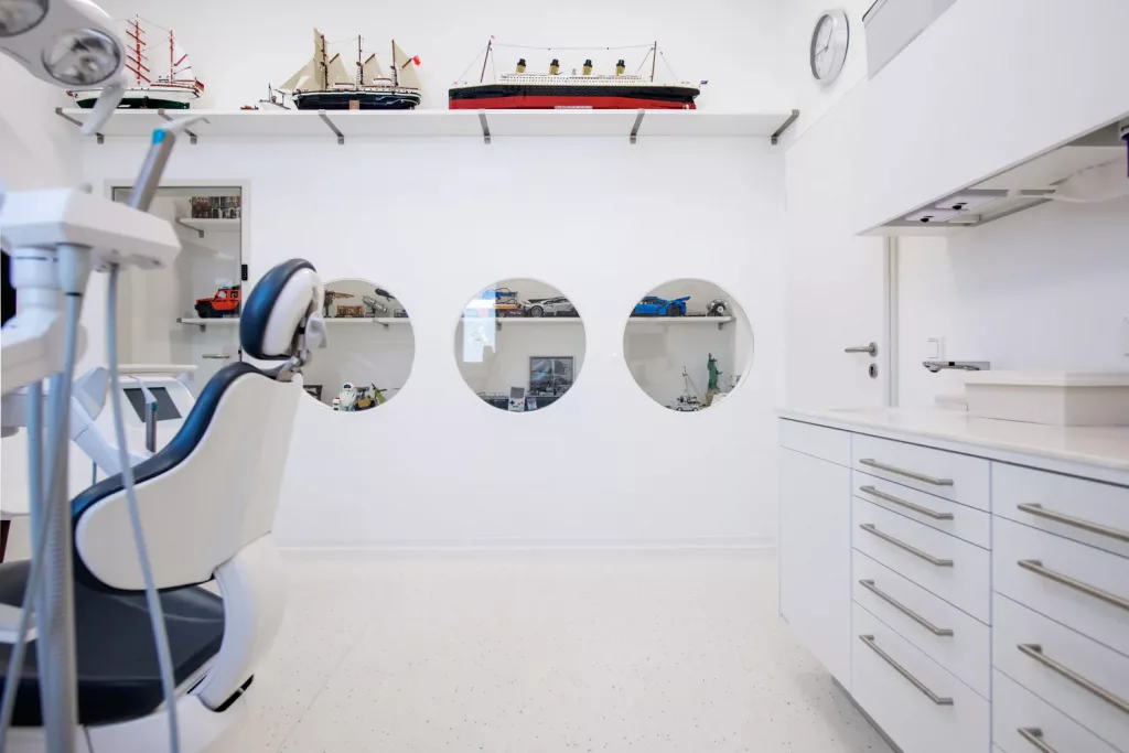 Loepraxis Dr Wilamowskiweb044 Dr Matthias Willamowski Kollegen Modern dental clinic interior with a reclining chair on the left and white cabinetry on the right decorative model ships on a shelf above and round viewing windows in the back wall