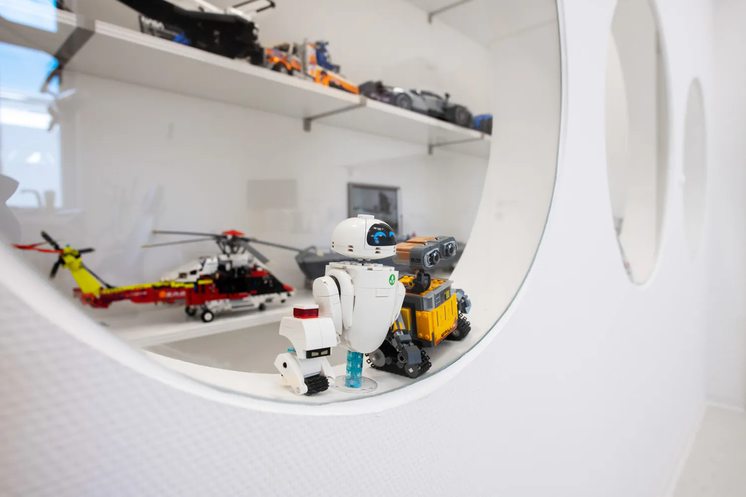 Weiße LEGO-Roboterfigur mit blauen Augen neben einem gelb-grauen Roboter auf einem geschwungenen weißen Regal, Modellfahrzeuge dahinter.