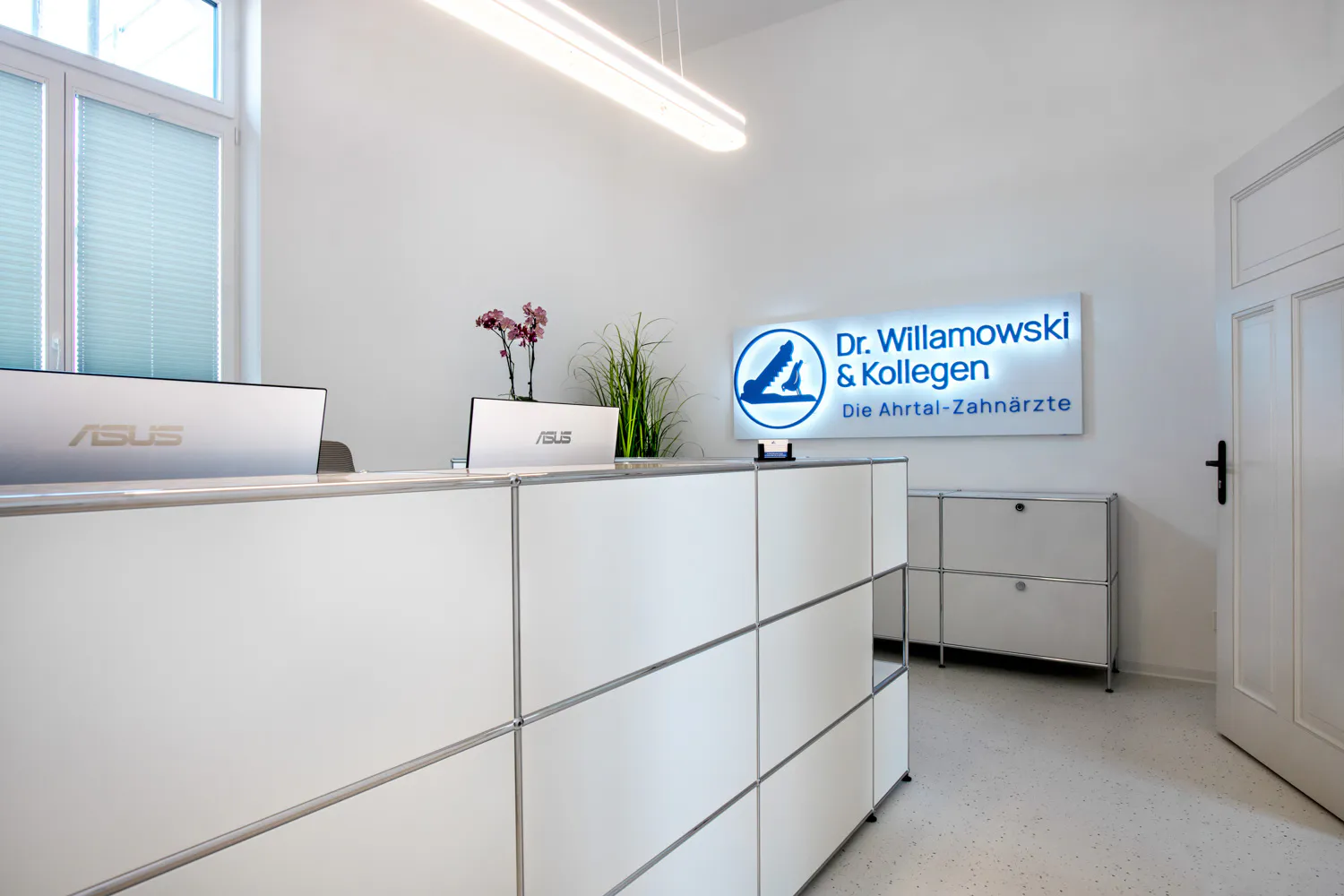 Empfang mit weißen Schränken, zwei Laptops und Topfpflanzen unter dem blau hinterleuchteten Schild „Dr. Willamowski & Kollegen“.
