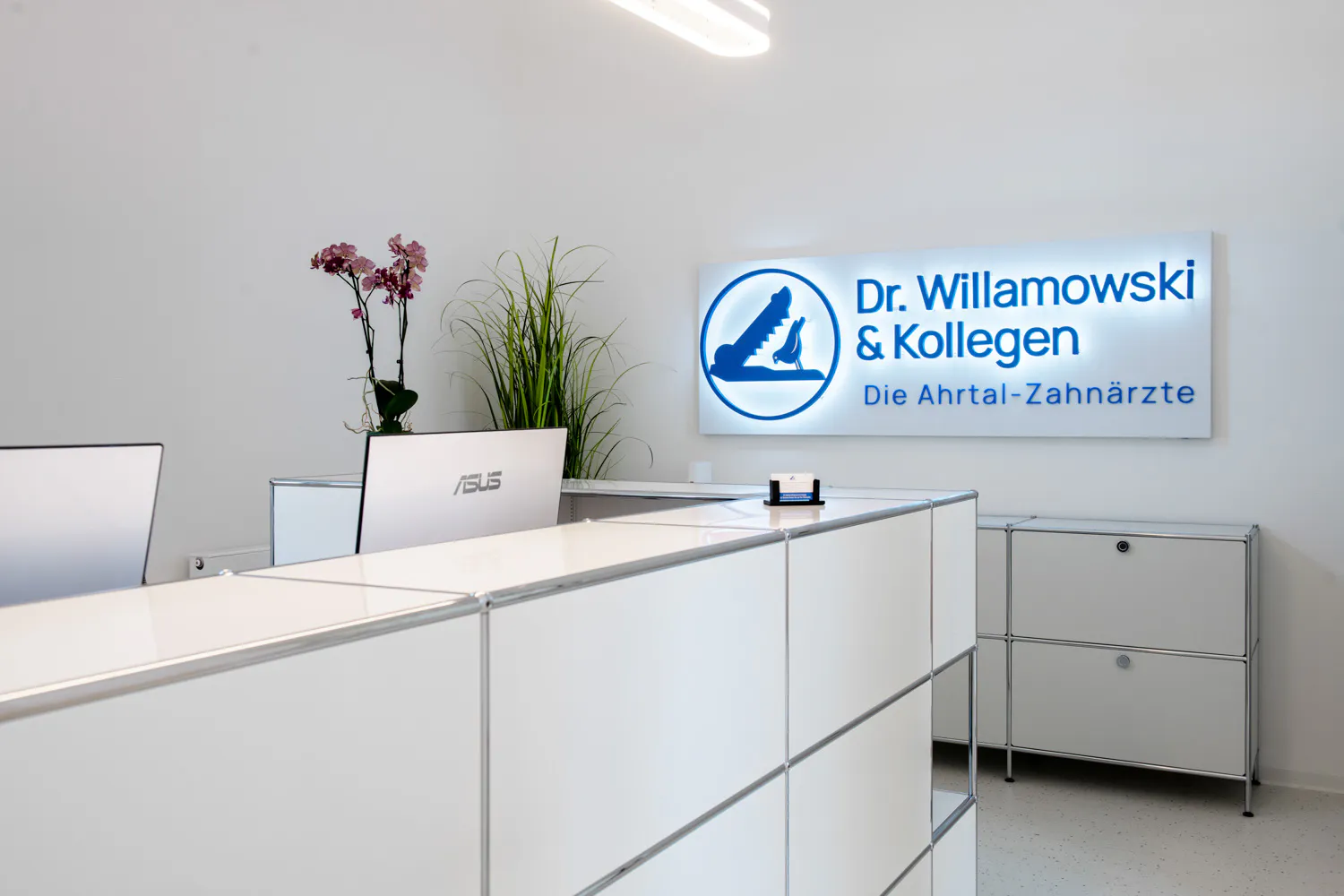 Loepraxis Dr Wilamowskiweb052 Dr Matthias Willamowski Kollegen Reception area of a dental clinic with white counters two laptops and potted plants plus a backlit sign reading Dr Willamowski Kollegen Die Ahltal Zahnarzte on the wall